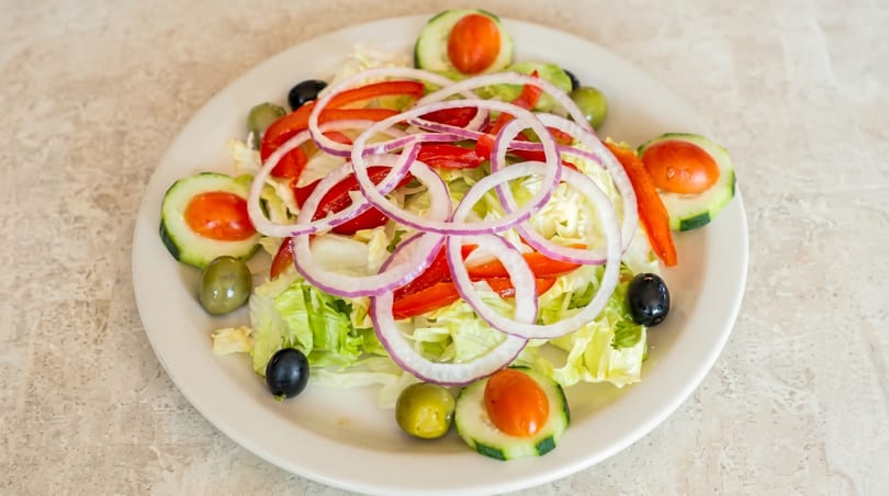 A fresh garden salad with lettuce, red onions, bell peppers, cucumbers, and olives on a white plate.