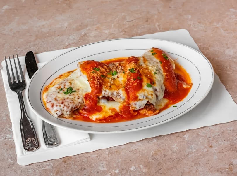 Chicken parmesan with melted mozzarella, red sauce, and parsley on a white oval plate with silverware.